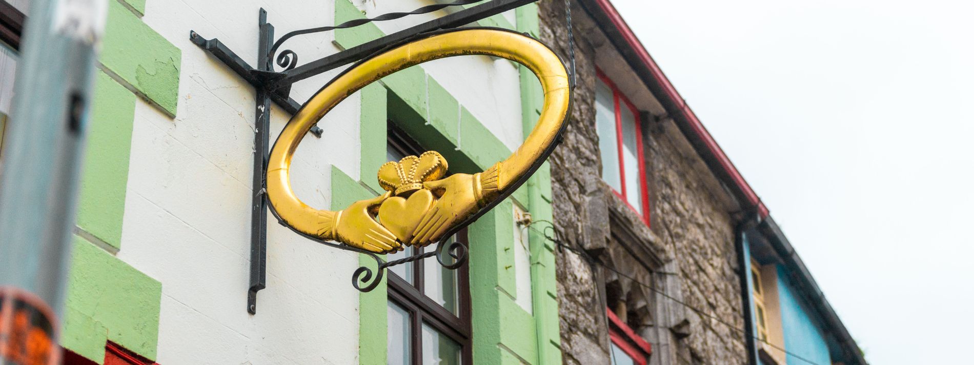 A golden ring with two hands holding a heart with a crown on the top as a hanging sign outside of a white and green old building in a historic area of Galway, Ireland, advertising Claddagh rings.