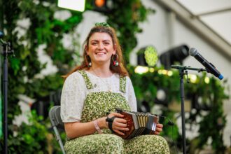 A smiling St. Patrick’s Day performer in floral green overalls playing a concertina, surrounded by festive foliage decoration.