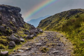 A rainbow stretches across the stony path to Carrauntoohil in County Kerry, Ireland, framed by rocky green ridges