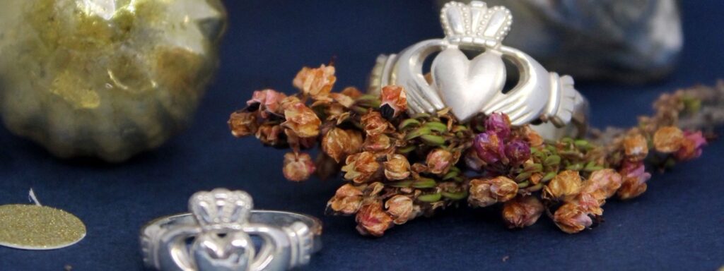 Two traditional claddagh rings made of silver rest on dried flowers. The Claddagh Rings show a pair of hands holding a heart