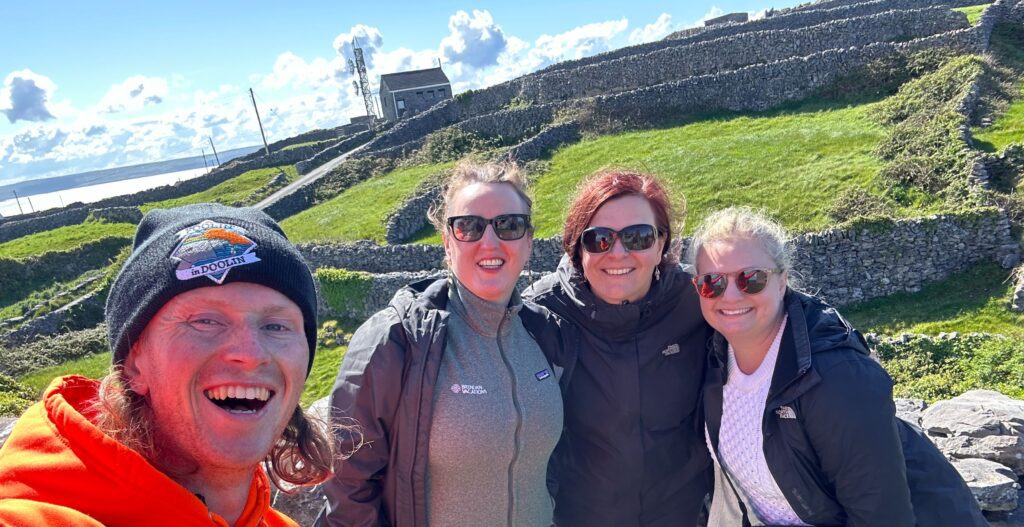 A man in a bright sweatshirt takes a selfie withe women wearing sunglasses in front of a green field bordered by stone walls in the Aran Islands in Ireland.