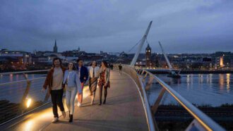 A group of people walking on a bridge at dusk with a cityb in the background in Derry, Ireland.