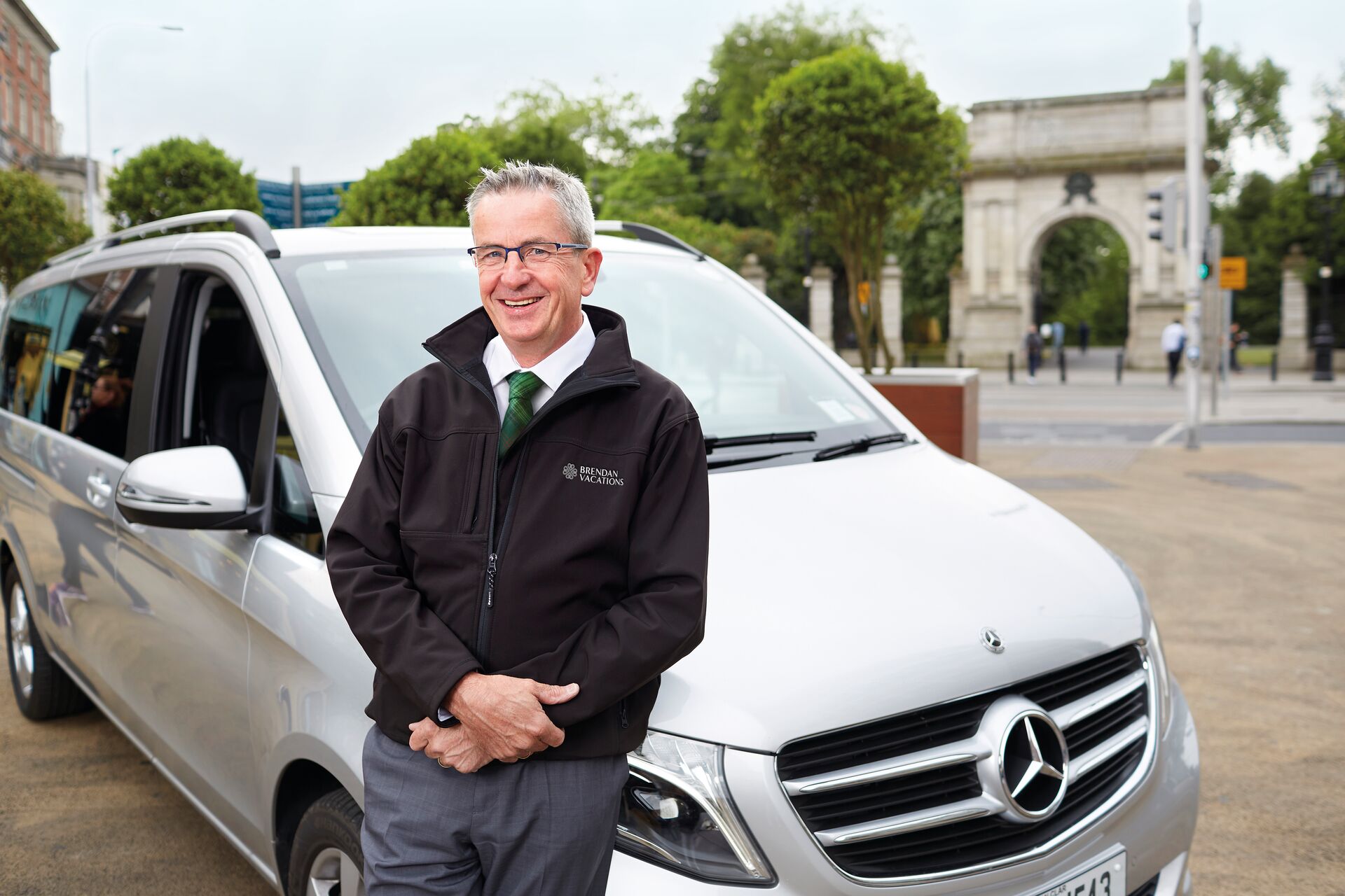 A smiling man in a black Brendan Vacations private chauffeur jacket stands in front of a car as part of the travel style "private chauffeur."