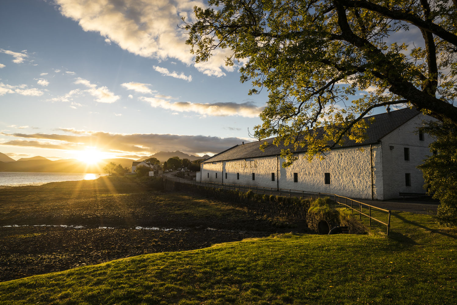 The sun is setting behind a white building with gray roof