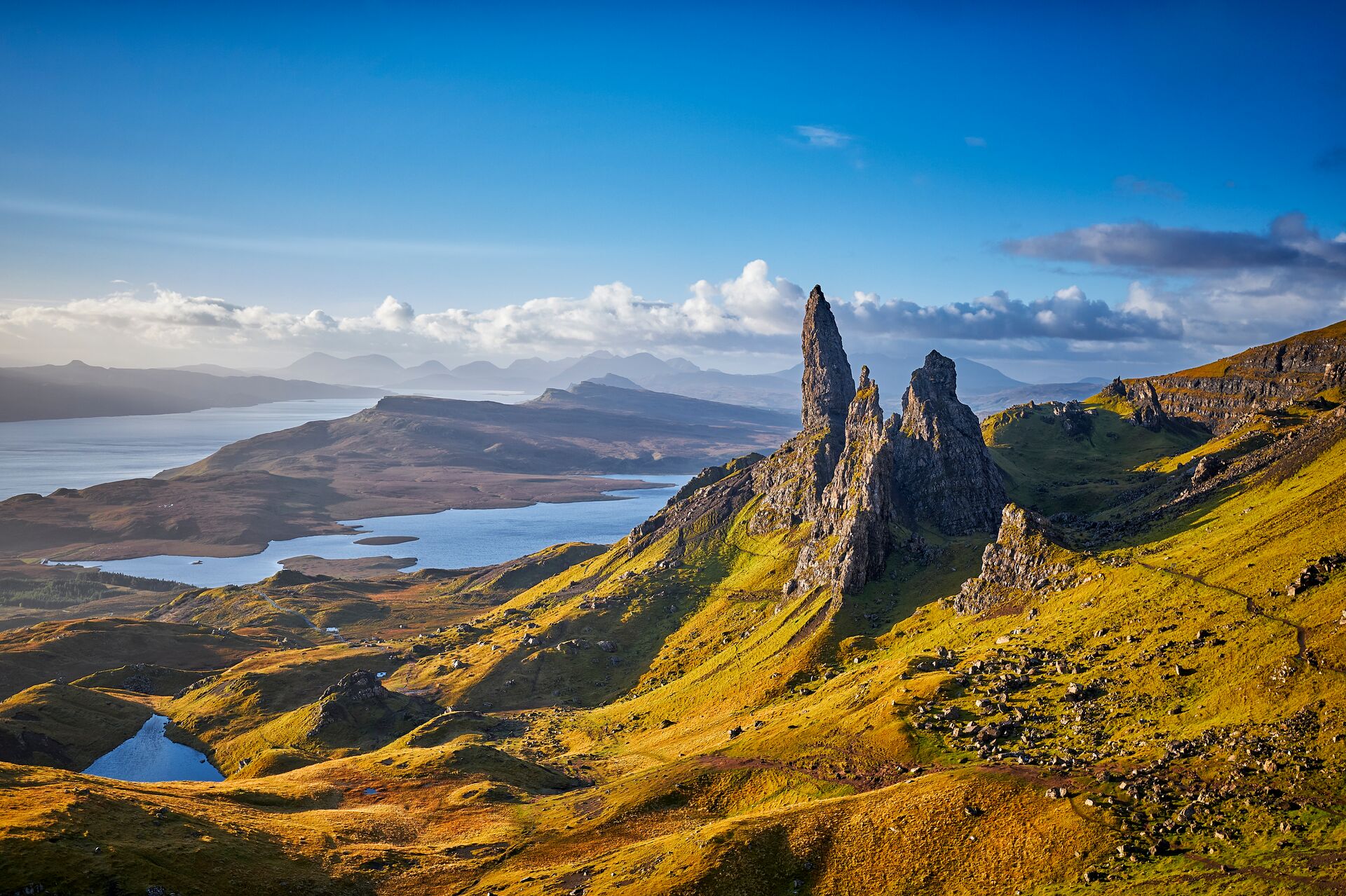 The tall crags of the Isle of Skye rise against a background of a blue sky overlooking lakes and mountains.