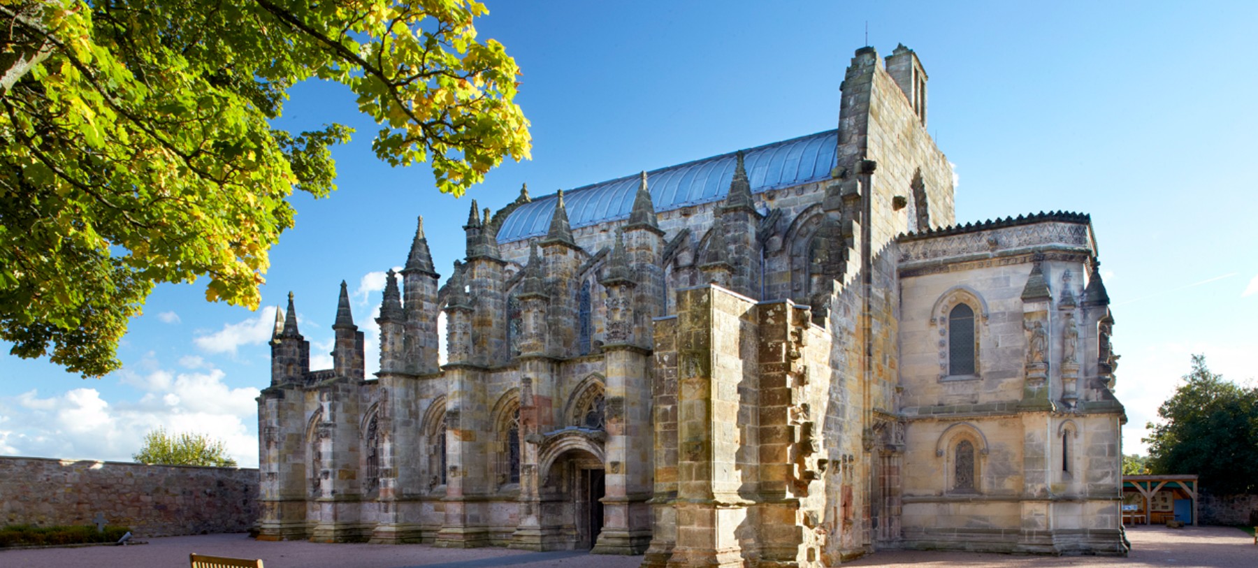 A white stone gothic building with five flying buttresses and a slate roof against a blue sky. The building is Rosslyn Chapel in Scotland.