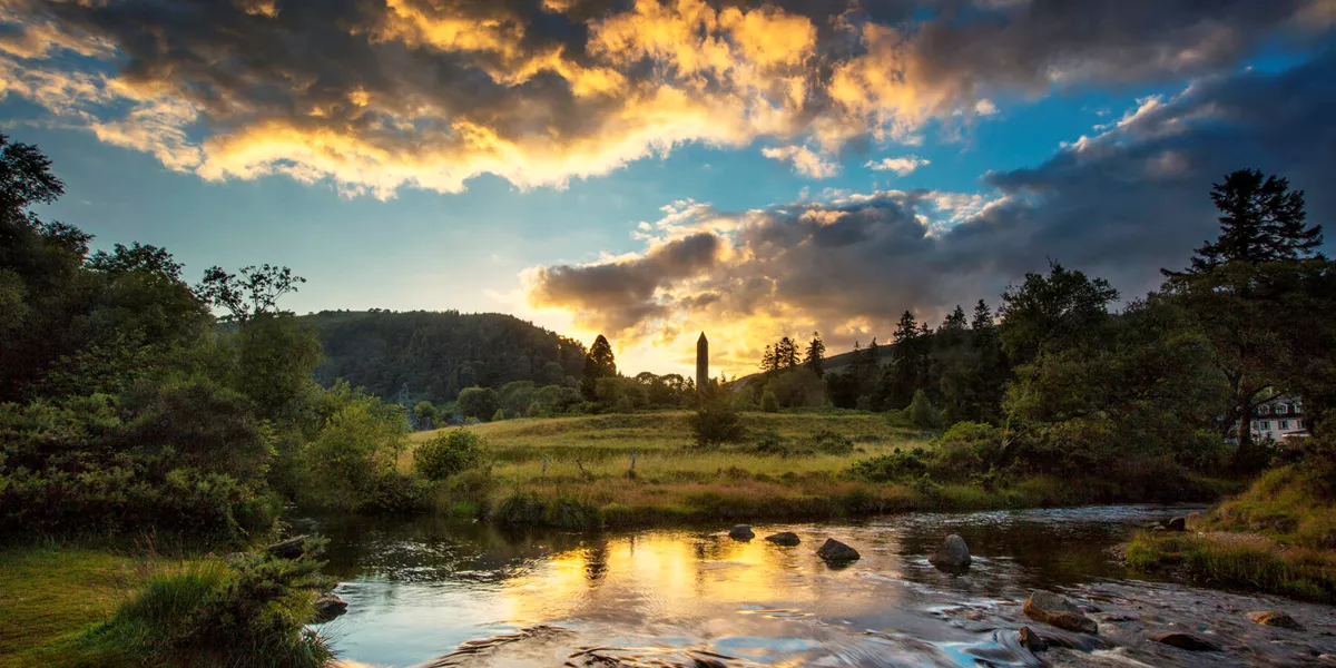 The sun rises over forested hills with a blue sky with clouds. In the foreground is a river and in the middle of a field there is a tower, flanked by trees.