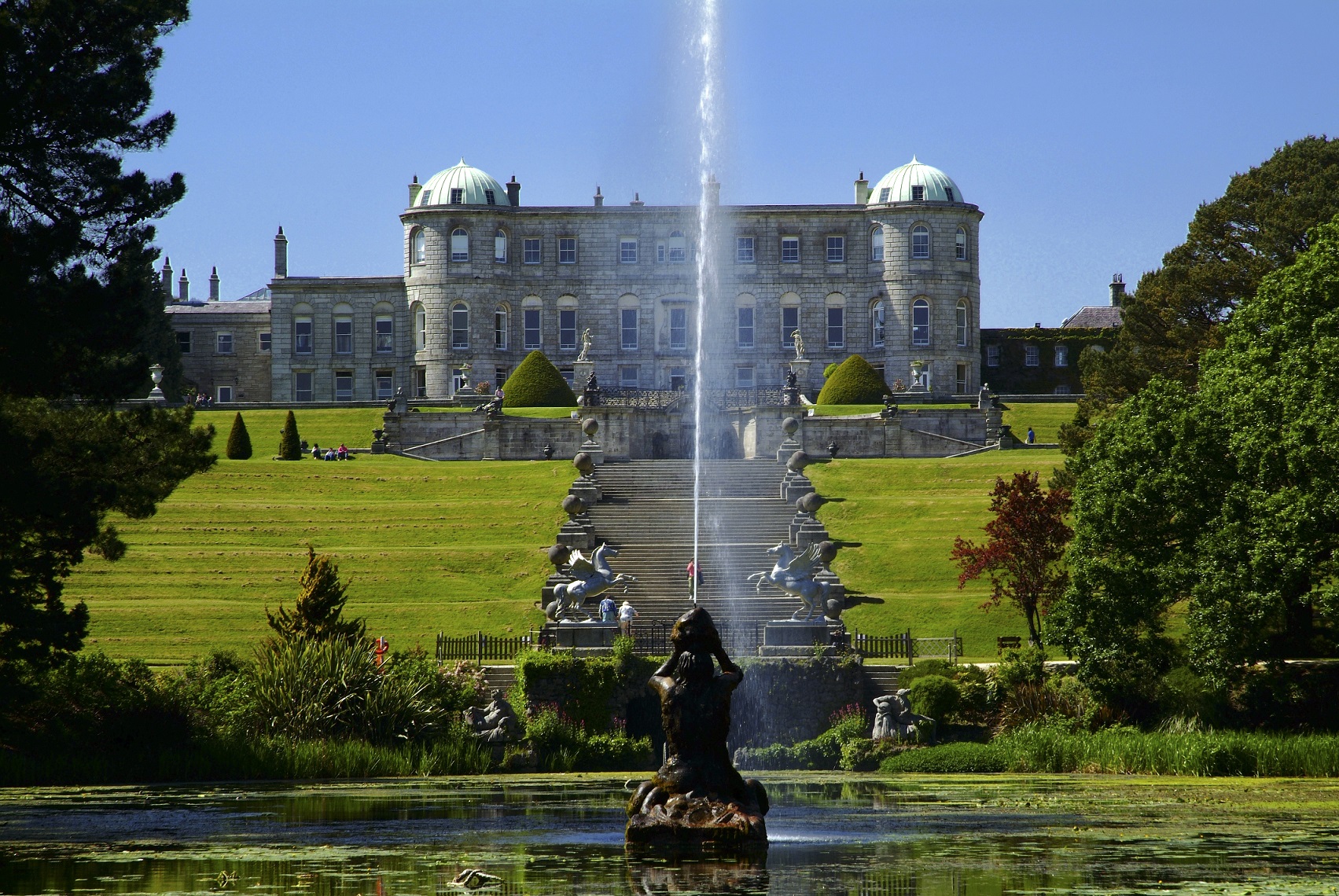A fountain in a lake shooting water in the air with a large house in the background