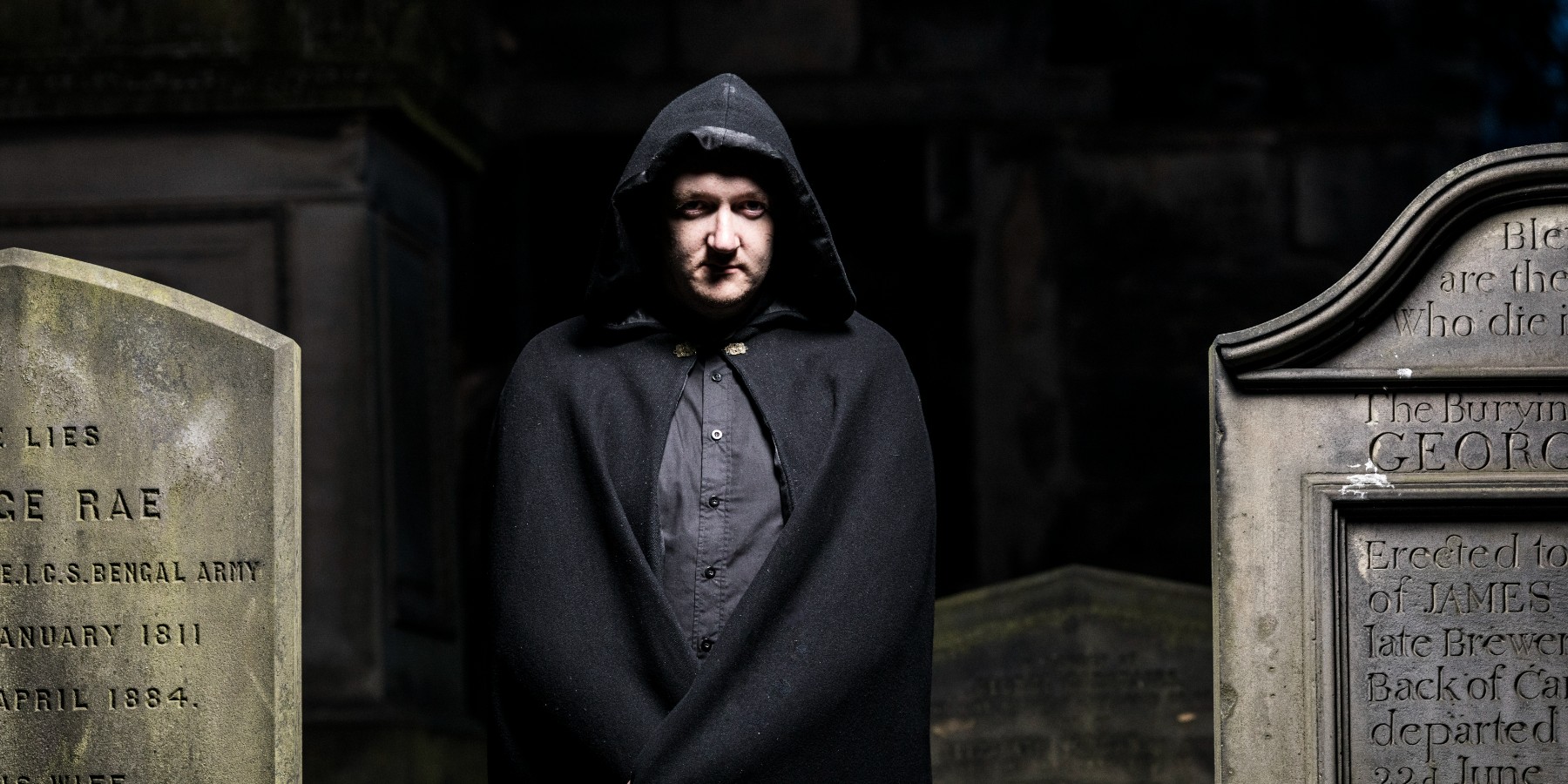 A man in a black cape with hood standing in a cemetery