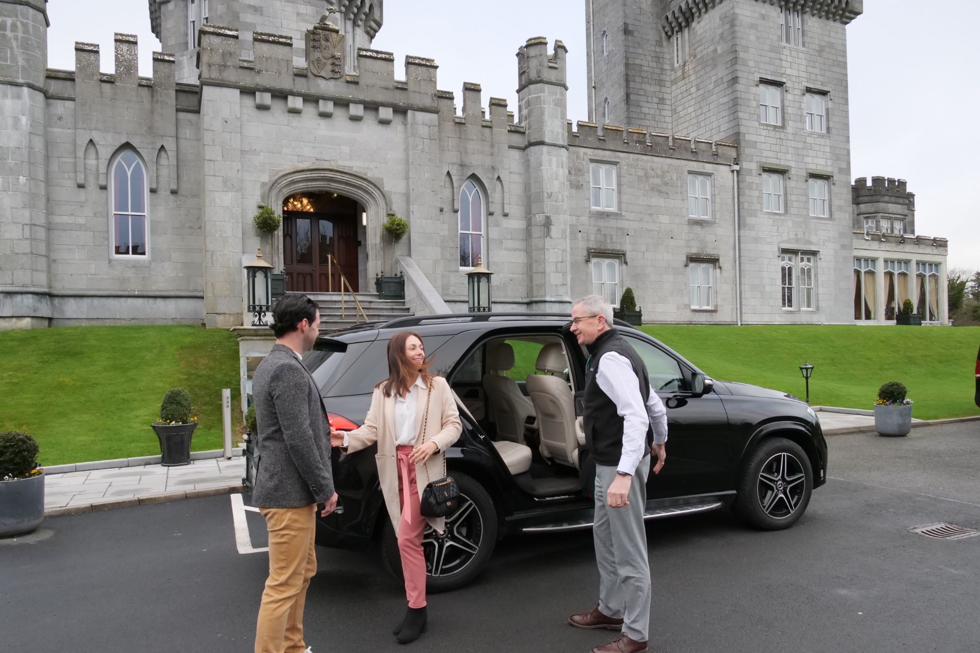 A private chauffeur holds open a car door as a male and female passenger exit the car in front of a large gray castle in Ireland.