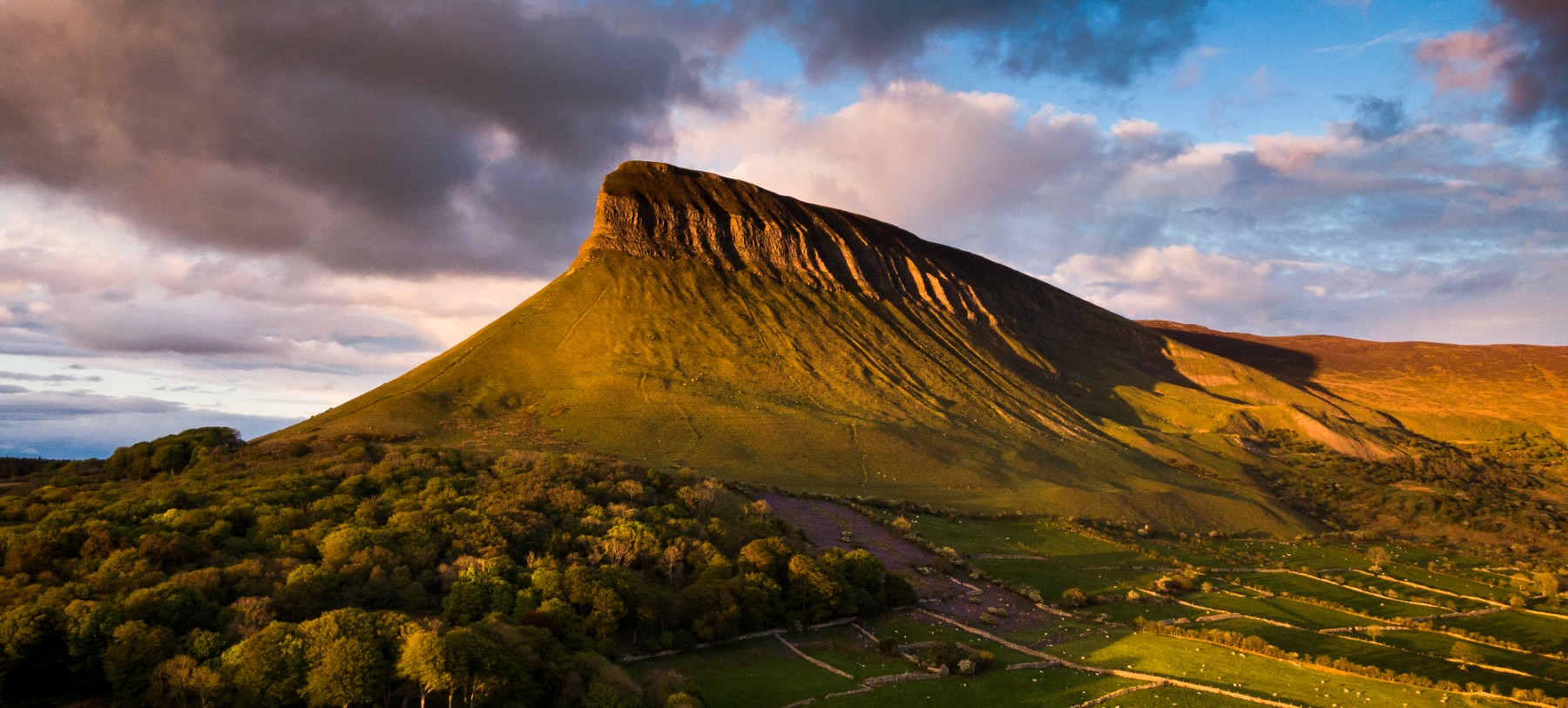 A large tabletop mountain called Benbulben rises against a blue sky with clouds in the background in the countryside of the west of Ireland, near Galway.