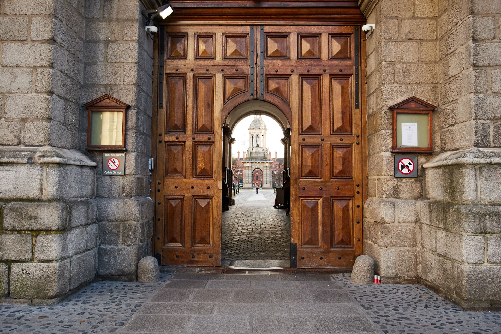 Large wooden doors in a grey stone wall with a walkway through the middle into the courtyard of Trinity College in Dublin, Ireland.