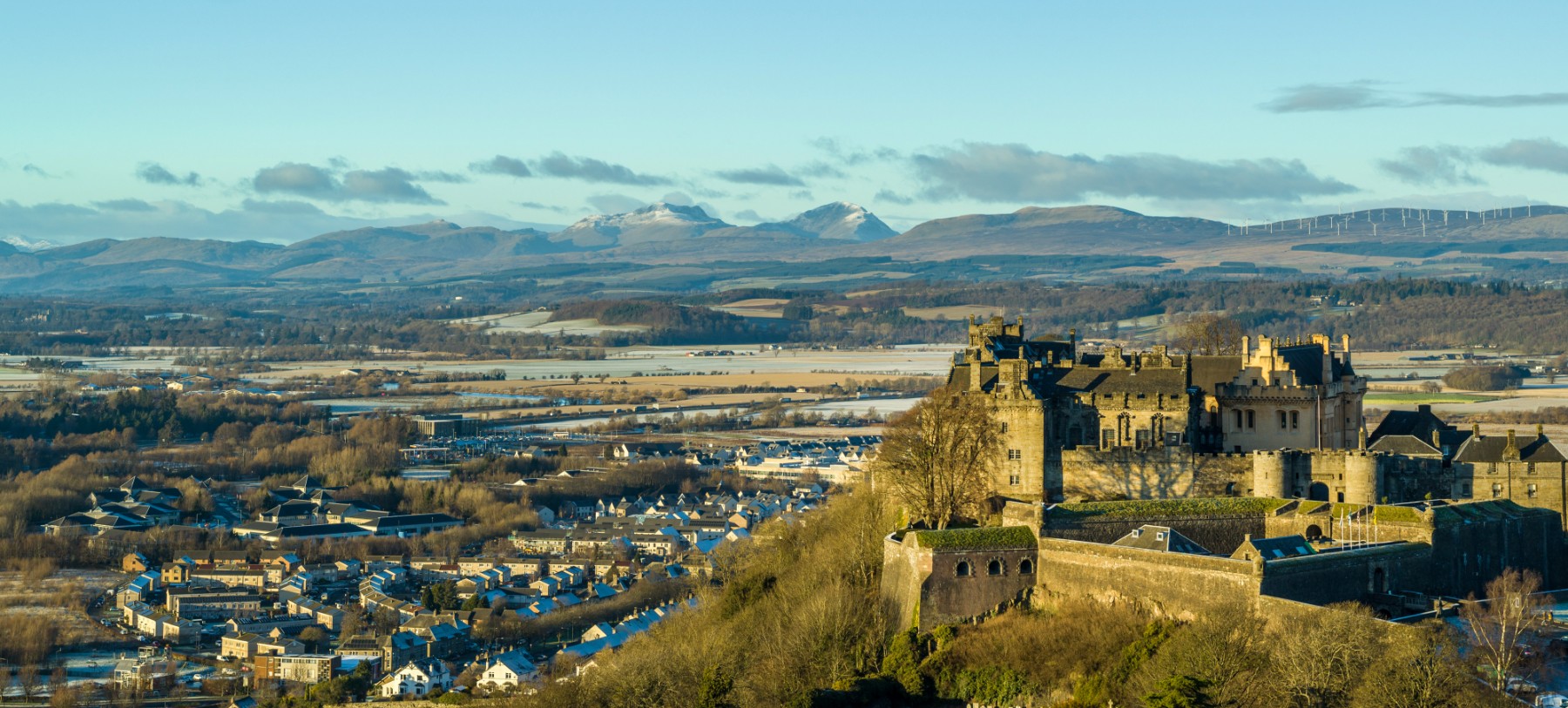 A stone manor home with a wall around it on a hill looks out over a town against a blue sky with clouds in Scotland.
