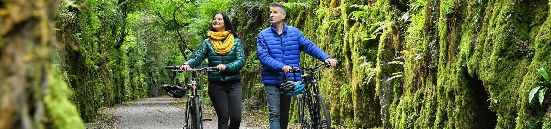 A man and woman walk their bikes in a pathway lined with moss and ferns covering walls in Ireland.