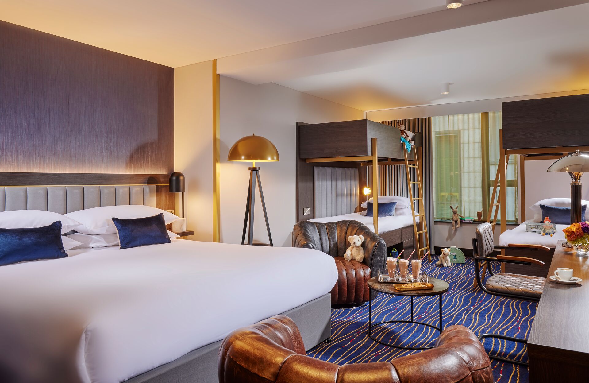 hotel room with one queen size bed, two bunk beds , two accent chairs with a teddy bear on one