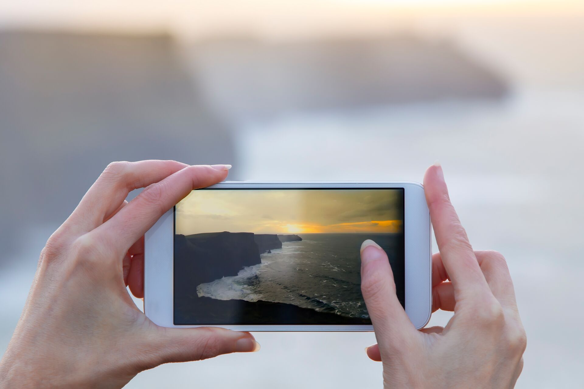 Hands holding a phone and taking a photo of Cliffs of Moher in ireland