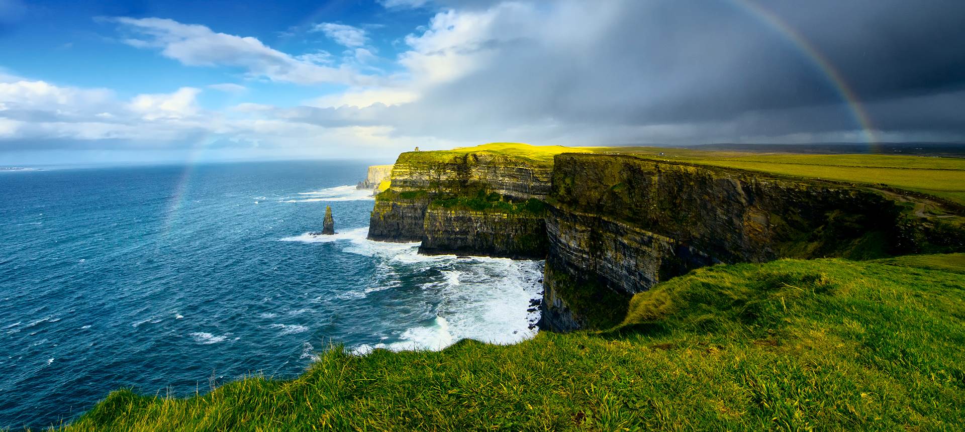 A rainbow shines over the ocean near a cliff 