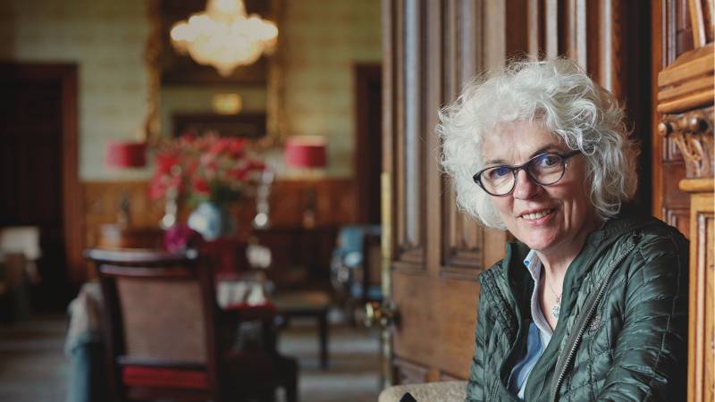 A woman with short curly white hair looks towards the camera, with a wooden door behind her and dining room in the background.