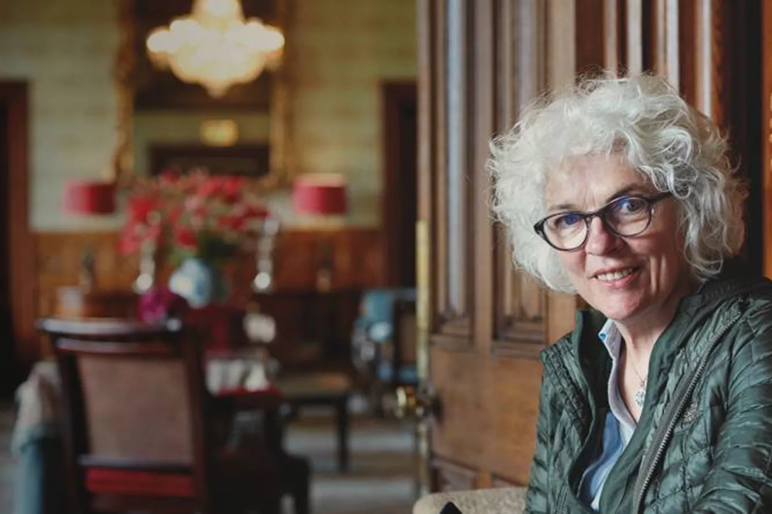 A woman with short curly white hair looks towards the camera, with a wooden door behind her and dining room in the background.