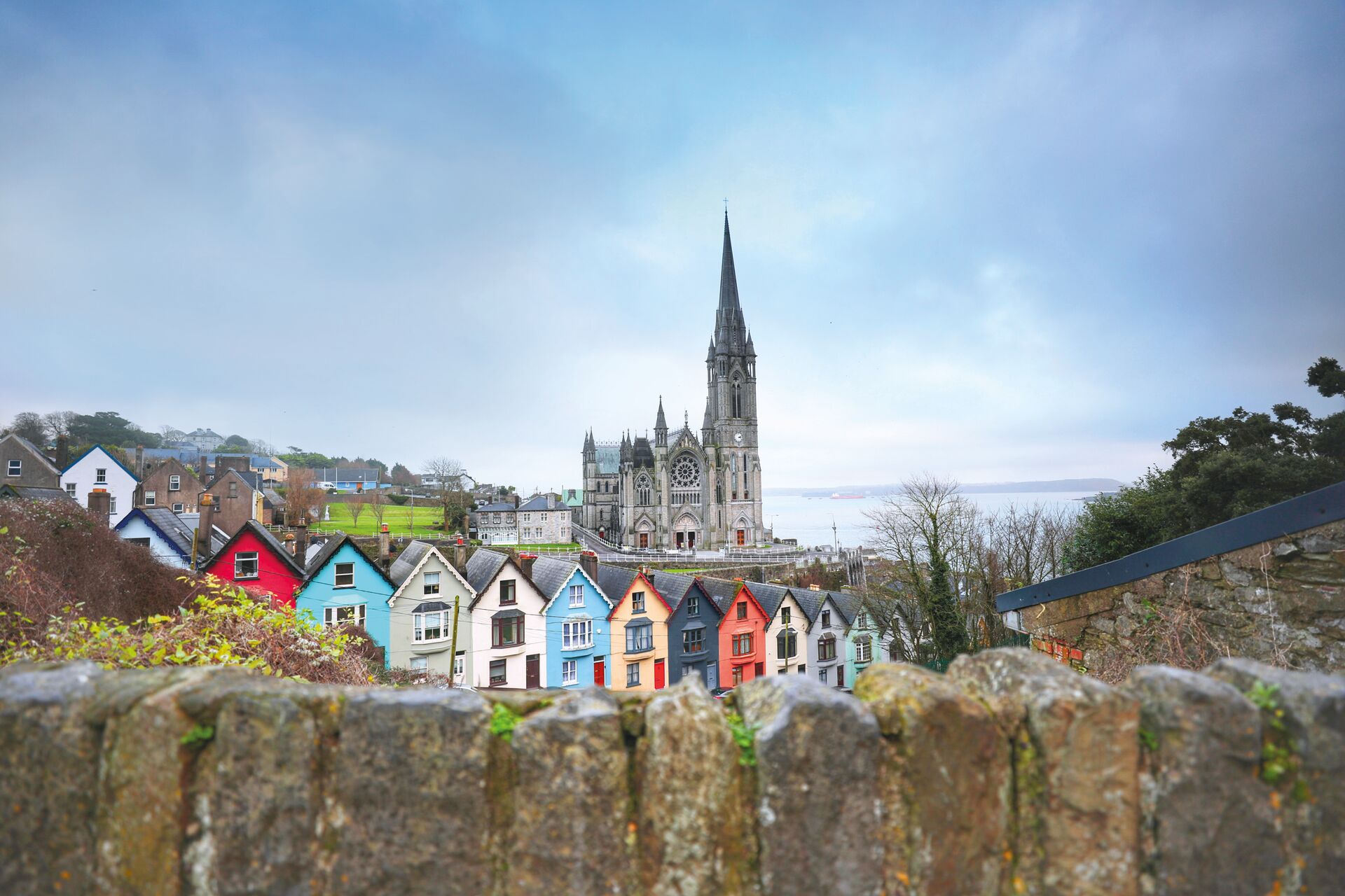 A view looking over a stone wall at a colorful row of houses in Cobh, county Cork, in the southeast of Ireland. A tall church steeple is in the background.